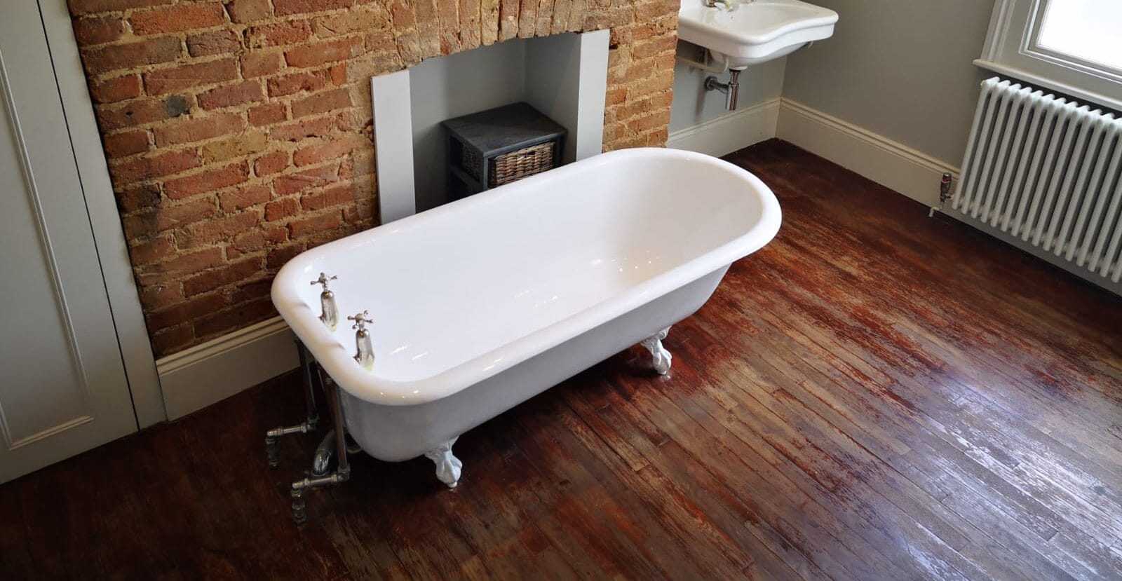 White roll top bath in front of a brick fireplace on a wooden floor